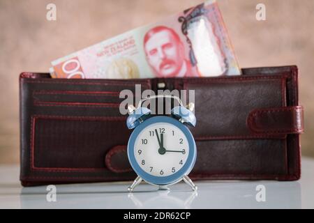 Stilvoller Wecker für fünf bis zwölf Personen. Im Hintergrund eine Brieftasche mit einem klebenden Haufen neuseeländischen Geldes. Hundert Dollarscheine Stockfoto