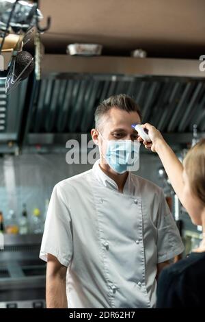 Zeitgenössische Koch in Uniform und Schutzmaske Blick auf Frau Mit einem Thermometer, das seine Körpertemperatur vor der Arbeit im misst Küche Stockfoto