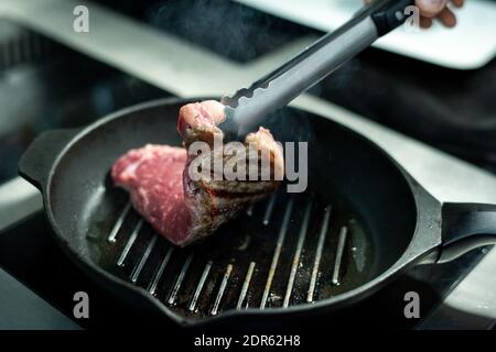 Koch mit Metallzange hält halb gerösteten Stück Fleisch über Heiße Grillpfanne mit Pflanzenöl beim Kochen Auf Elektroherd Stockfoto