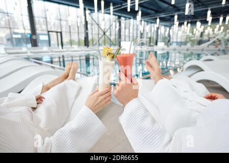 Hände von jungen Mann und Frau in weißen Bademänteln entspannend Auf Liegestühlen vor der Kamera im Freizeitzentrum und Klirrend mit Cocktails Stockfoto