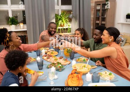 Glückliche Familie von jungen afrikanischen Paar, zwei Teenager und kleinen Jungen und Mädchen klirren mit Gläsern hausgemachten Rotwein über serviert festlichen Tisch Stockfoto