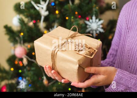 Womans Hände halten schöne Geschenk in Bastelpapier eingewickelt, Weihnachtsbaum auf dem Hintergrund, Zero Waste Konzept Stockfoto