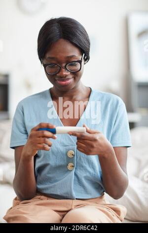 Vertikales Porträt einer glücklichen afroamerikanischen Frau, die den Schwangerschaftstest betrachtet Während Sie zu Hause auf dem Bett sitzen Stockfoto
