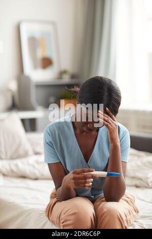 Vertikales Porträt einer besorgten afroamerikanischen Frau, die einen Schwangerschaftstest ansieht Während Sie zu Hause auf dem Bett sitzen Stockfoto