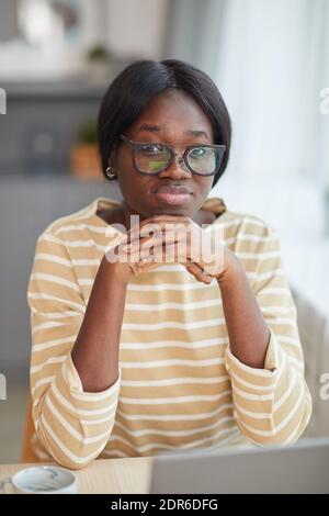 Vertikales Porträt einer jungen afroamerikanischen Frau, die eine Brille trägt und schaut Bei der Kamera, während Sie am Schreibtisch in gemütlicher Wohneinrichtung sitzen Stockfoto