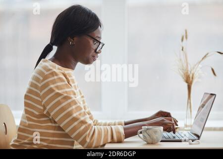 Seitenansicht Porträt der jungen afroamerikanischen Frau mit Laptop, während Sitzen am Tisch in gemütlichen Innenraum Stockfoto