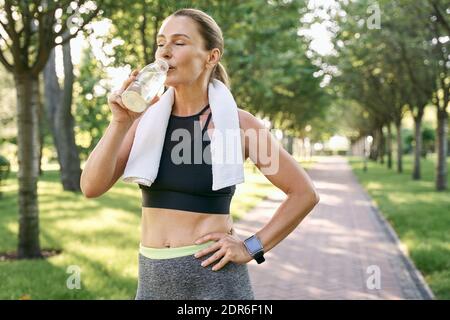 Bleib hydratisiert. Müde fit Frau in Sportbekleidung erfrischend, Trinkwasser nach dem Joggen in einem grünen Park an einem sonnigen Tag Stockfoto