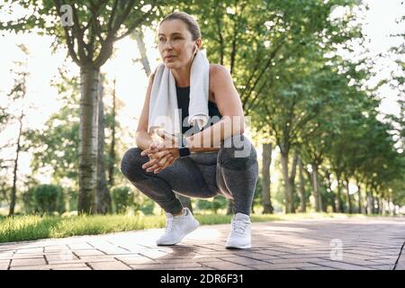 Werden Sie stärker. Müde sportliche Frau mittleren Alters in Sportkleidung mit Handtuch um den Hals ruhen nach dem Laufen in einem grünen Park an einem sonnigen Tag Stockfoto