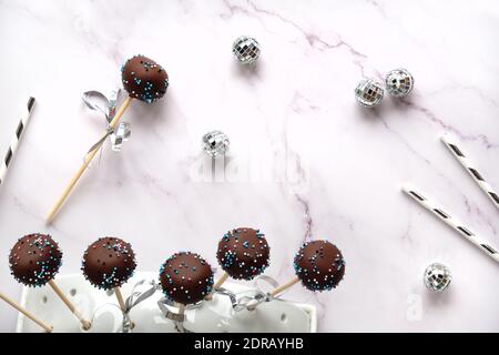 Leckere Kuchen Pops mit Schokolade und farbigen Zucker Glasur bedeckt. Party-Dekorationen, Trinkhalme aus Papier und Diskokugeln auf weißem Marmorhintergrund. Merr Stockfoto