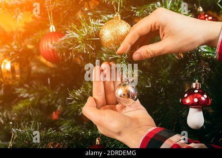 Nahaufnahme einer Frau in einem karierten Hemd hängt eine schöne glänzende Goldkugel auf einem Weihnachtsbaum auf einem hölzernen Wandhintergrund. Weihnachten und Neujahr Home PR Stockfoto