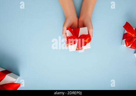 Kinderhände halten eine weiße Geschenkbox mit einer roten Schleife auf blauem Hintergrund. Urlaub Geschenk Konzept.Flat Lay Stockfoto