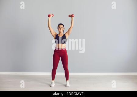 Athletische Frau, die mit erhöhten Hanteln in ihren Händen steht und ihren straffen athletischen Körper zeigt. Stockfoto