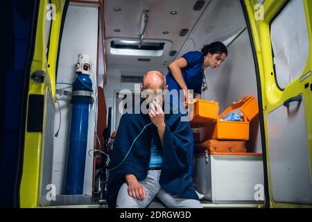 Mann sitzt mit Sauerstoffmaske, Krankenschwester in Uniform verpackt Ausrüstung in medizinische Tasche. Stockfoto