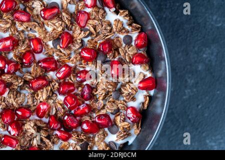 Granola mit Haferflocken, getrockneten Früchten, Honig, Rosinen, Schokoladenstückchen, Nüssen und roten Granatapfelkernen mit Joghurt in einer schwarzen Schüssel, Nahaufnahme, Draufsicht. Er Stockfoto