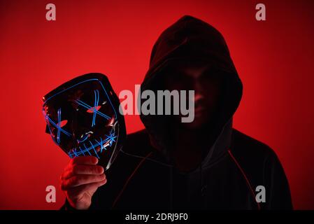 Mann im Hoodie mit verschwommenem Gesicht hält Neon glühende Gruselmaske. Anonymes Konzept Stockfoto