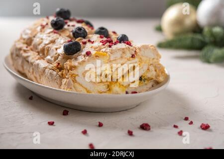 Hausgemachte Meringue-Rollkuchen mit Sahne, Beeren und Mandel auf dem Teller, selektiver Fokus, im Hintergrund Weihnachtsbaum Äste mit Spielzeug Stockfoto