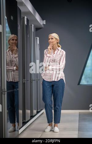 Frau im gestreiften Hemd Auswahl Kühlschrank in einem Showroom und Sieht nachdenklich aus Stockfoto