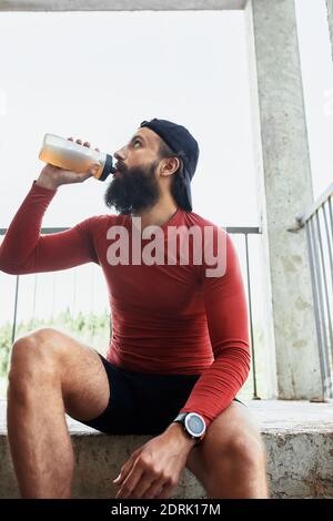 Seitenansicht des aktiven bärtigen Sportlers, der Wasser aus der Glasflasche trinkt, während er sich ausruhte und an einem hellen Tag auf der Treppe saß. Stockfoto