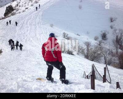 Kerl in einer roten Jacke steht auf einem Snowboard auf einem Berggipfel im Schnee, Blick von hinten Stockfoto