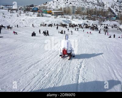 Mädchen in einem weißen Trainingsanzug reitet einen Schlitten auf Schnee von einem Berg auf den Grund an einem sonnigen Tag, Blick von hinten Stockfoto