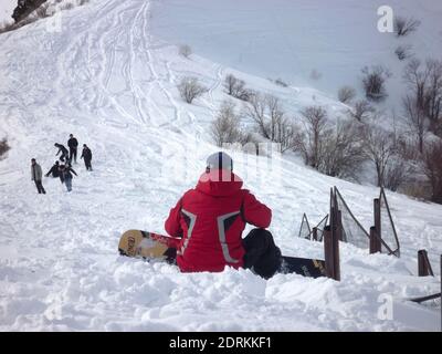 Kerl mit einem Snowboard in einer roten Jacke sitzt im Schnee auf der Spitze eines Berges, ein Blick von hinten Stockfoto