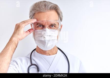 Arzt Mann trägt Schutzbrille für die Vorbereitung auf die Arbeit, ältere Mann in weißem medizinischen Anzug schaut auf Kamera isoliert auf weißem Hintergrund Stockfoto