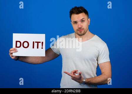 Schöner stilvoller Mann, der mit einem Schild in der Hand im Studio auf blauem Hintergrund steht. Wort tun es auf weißem Blatt Papier. Stockfoto