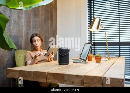 Frau, die mit dem intelligenten Lautsprecher spricht, während sie am digitalen Tablet im gemütlichen Heimbüro arbeitet. Konzept eines Smart Home und Künstliche Intelligenz Stockfoto