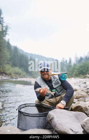 Lächelnder Fischer in legerer Kleidung, der seine Rute im Mund und Forelle in den Händen hält. Glücklicher Mann, der auf Steinen zwischen dem Gebirgsfluss sitzt. Stockfoto