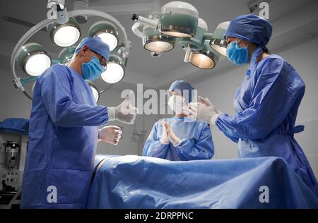 Gruppe von Ärzten in sterilen Handschuhen und schützenden Gesichtsmasken stehen beim Patienten nach erfolgreicher Operation. Männlicher plastischer Chirurg tut Daumen nach oben Geste, während Assistenten applaudieren. Stockfoto