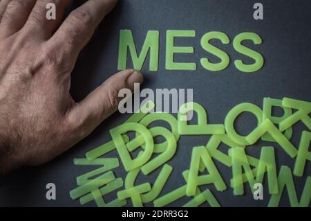 Menschliche Hand neben dem Wort Mess mit Plastikbuchstaben auf grauem Papier Hintergrund geschrieben, mit einem Haufen von Buchstaben in Unordnung, Konzept Stockfoto