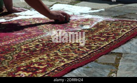 Eine Frau putzt einen schmutzigen Teppich auf der Straße, im Hof des Hauses aus der Nähe. Mit sauberem Wasser abspülen. Viel Schaum aus dem Pulver Stockfoto