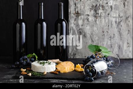 Weinflaschen Käsenüsse und Trauben auf Schiefertafel. Snacks an der Weinbar. Weinkomposition auf dunklem rustikalem Betonhintergrund mit Kopierraum Stockfoto