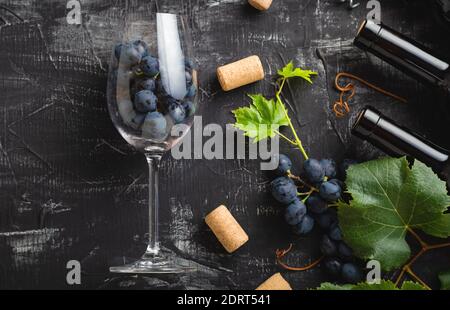 Weinglas voller Trauben im Inneren. Weinflaschen, Weintrauben mit Blättern und Weinreben Weinkorken auf dunklem rustikalem Betonhintergrund. Flacher Lay-Wein Stockfoto