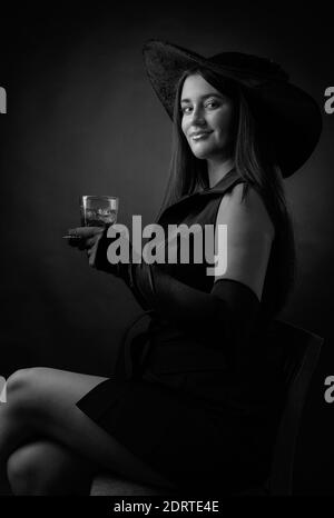 Attraktive Mädchen mit alkoholischen Getränk in gefrorenem Glas. Schöne Brünette in schwarzem Hut hält Glas mit kalten Cocktail. Schwarz und Weiß. Stockfoto