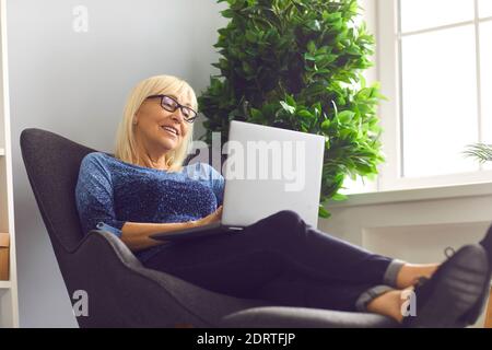 Reife Frau mit Brille sitzt in einem Sessel und arbeitet zu Hause mit einem Laptop. Stockfoto