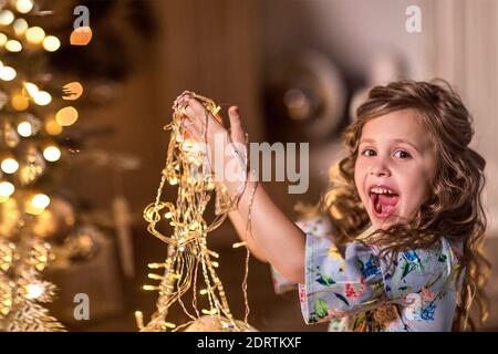 Portrait eines schönen lachenden Mädchens mit lockigen langen Haaren auf einem Hintergrund von verschwommenen Weihnachtslichtern (Bokeh) und einem Weihnachtsbaum. Das Konzept des wi Stockfoto