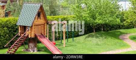 Kleine Holzblockhaus Hütte mit Treppenleiter und Holzrutsche auf Kinderspielplatz im Park oder Hof. Panoramablick. Grüner Rasen Rasen Garten Stockfoto