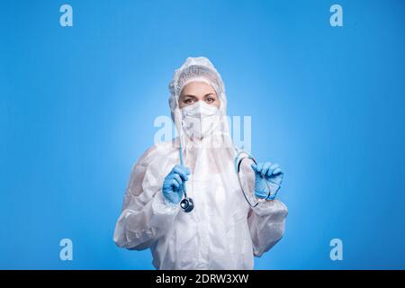 Weibliche Sanitäter tragen Schutzanzug und halten Stethoskop auf blauem Hintergrund. Arzt behandelt Atemwegserkrankungen im Krankenhaus Stockfoto