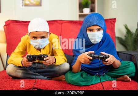 Zwei muslimische Kinder mit medizinischer Gesichtsmaske beschäftigt spielen Video Spiel mit Gamepad zu Hause - Konzept der Kinder auf Spiel während Coronavirus covid-19 Lockdown Stockfoto