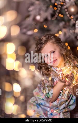 Portrait eines schönen lachenden Mädchens mit lockigen langen Haaren auf einem Hintergrund von verschwommenen Weihnachtslichtern (Bokeh) und einem Weihnachtsbaum. Das Konzept des wi Stockfoto