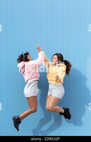 Zwei freundliche Mädchen springen und klatschen auf blauem Grund in die Luft. Leerzeichen für Text. Freundschaftskonzept. Stockfoto