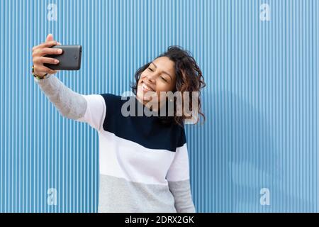 Junge und lächelnde lateinerin, die mit ihrem Mobiltelefon auf blauem Hintergrund ein Porträt zeichnet. Leerzeichen für Text. Stockfoto