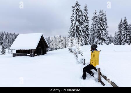 Frau sitzt auf einem Holzzaun an einer traditionellen Holzhütte in schneebedeckten malerischen Almwiese in den Julischen alpen im Winter, Pokljuka, Slowenien Stockfoto