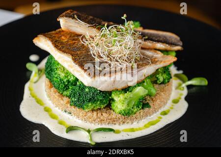 Seebarschfilet mit Brokkoli und Quinoa auf schwarzem Teller. Restaurant, das Essen serviert. Gesunde Ernährung Konzept Stockfoto