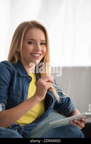 Glücklich blonde Frau Blick auf die Kamera, während halten digitale Tablette Und Brillen zu Hause Stockfoto