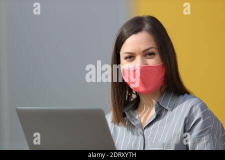 Zufriedene Frau trägt Schutzmaske und mit Laptop Blick auf Kamera auf der Straße Stockfoto