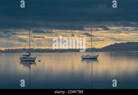 Zwei Segelboote mit Reflektionen am Anker im Christchurch Hafen mit Mudeford Beach Hütten im Hintergrund an EINEM ruhigen, stillen Tag bei Sonnenaufgang Stockfoto