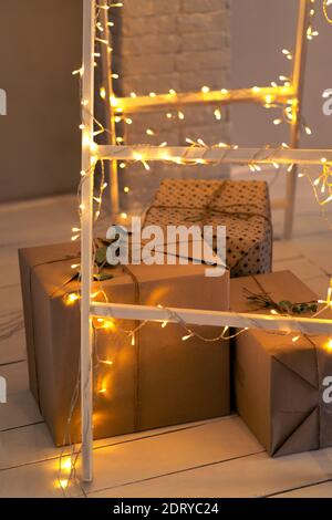 Geschenke unter der Treppe mit Girlande. Festlicher Hintergrund mit Lichtern. Große Schachteln in Kraftpapier Neujahrsgeschenke im Innenraum verpackt. Stockfoto