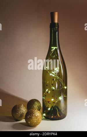 Girlande in Glasflasche für Wein und drei goldglänzende weihnachtskugeln. Selektiver Fokus, vertikale Ansicht Stockfoto